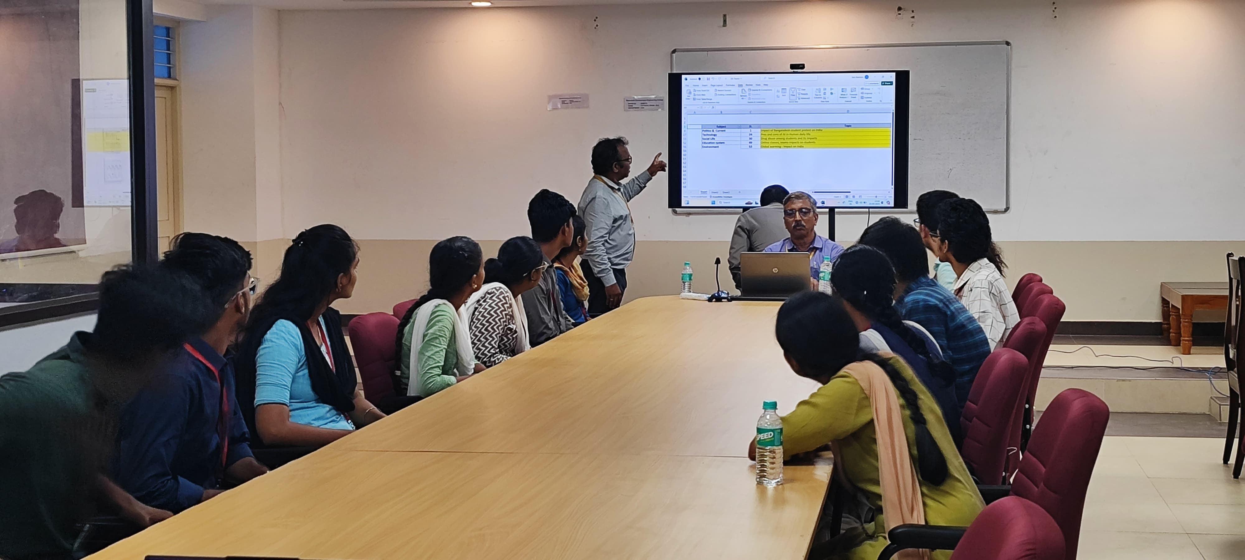Students seated along a long boardroom table watching a facilitator present at a screen