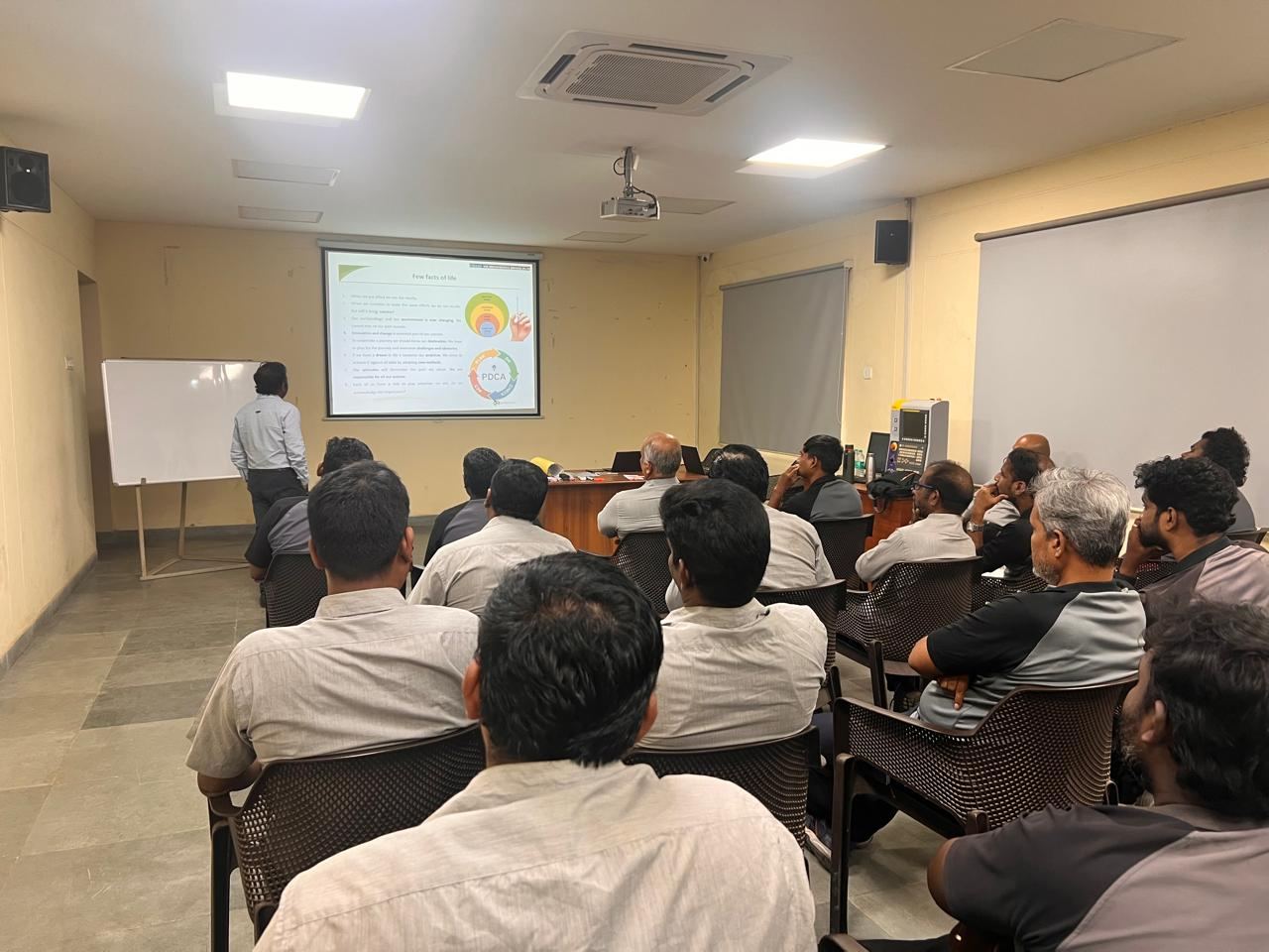 A room of seated participants facing a facilitator presenting beside a projected slide